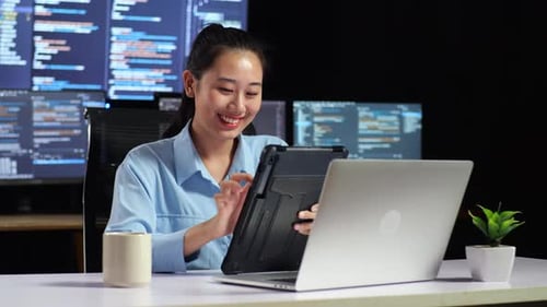 Asian Female Programmer Using Tablet While Writing Code By A Laptop Using Multiple Monitors