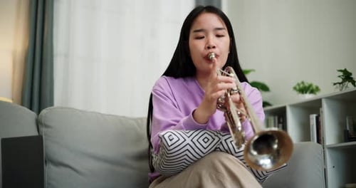 A young woman practices playing the trumpet while sitting on a sofa in a cozy living room.