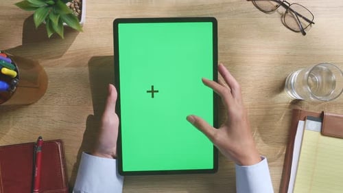 Top down view of man's hands using a green screen tablet on a wooden desk
