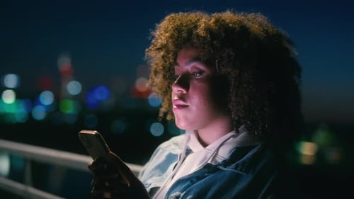 Black woman standing on the bridge at night and browsing her mobile phone. Shot with RED helium came