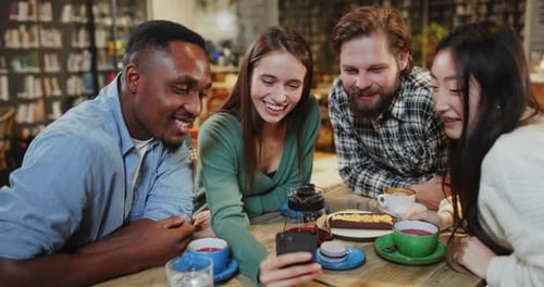 Diverse Multicultural Joyful Friends Sharing Table in Coffee Shop Watching Funny Videos Using