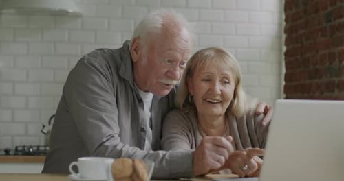 Elderly Couple Man and Woman Talking and Looking at Laptop Screen Discussing Content in Kitchen at