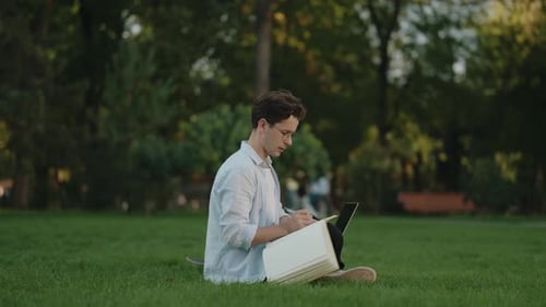 Young Man Working in Park on Grass with Notebook and Tablet