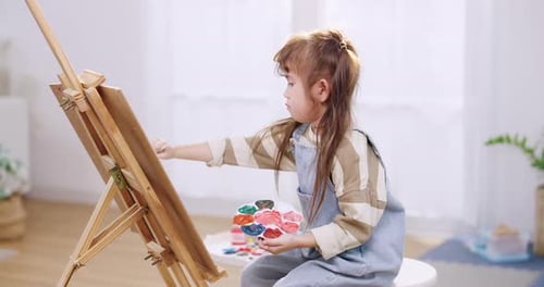 Asian preschooler daughter paints on easel canvas, creative workshop for kids at home, fine motor