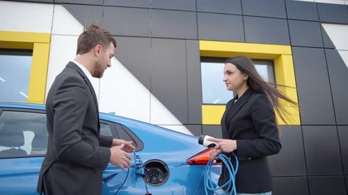 Businessman in a dealership shows a girl how to charge an electric car at a charging station