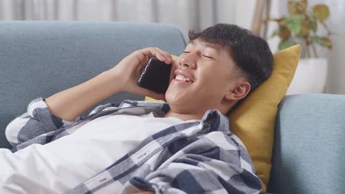 Close Up Of Asian Teen Boy Talking On Smartphone While Lying On Sofa In The Living Room At Home