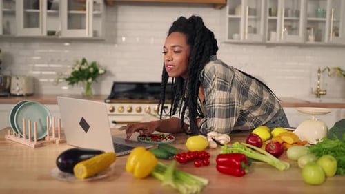 Young Woman Eats Salad at Laptop in Kitchen
