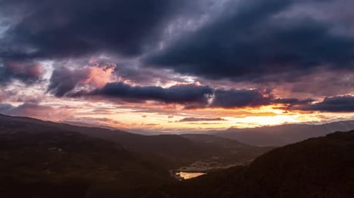 Dark clouds whirl in the sunset sky backlit by the orange sun. Rolling hills stretch to the horizon.