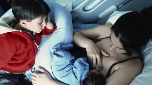 Mother breastfeeding newborn in hospital bed, with older sibling in red hoodie watching closely,