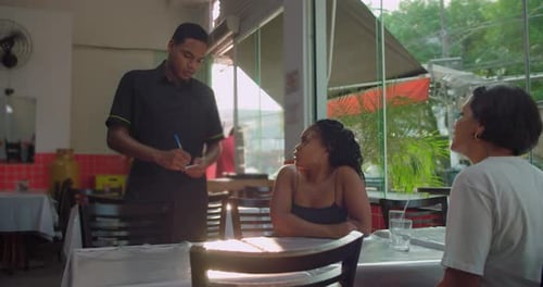 Latin waiter of African descent takes order from two women seated at restaurant table, friendly