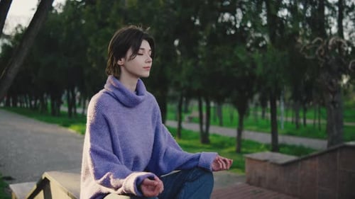 Young Woman Sits on a Bench in a Park Practicing Meditation with Eyes Closed During Late Afternoon