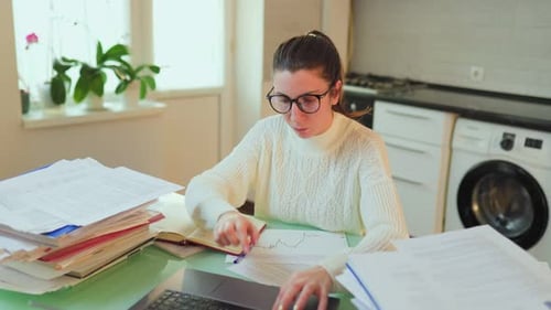 A Student with Glasses in Elearning Surrounded By Documents While Working on a Laptop at Home