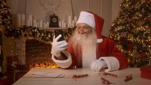 Santa Claus is Using the Phone Sitting at a Table Near a Christmas Tree and Decorated Fireplace