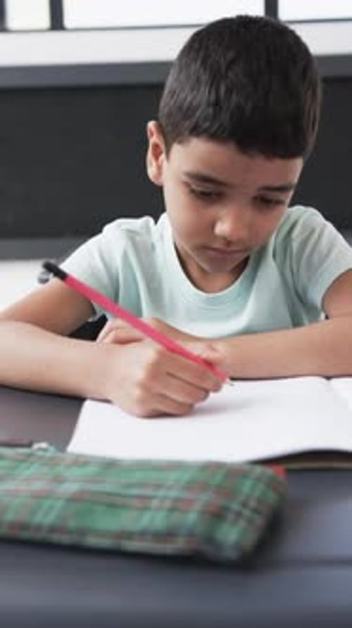 Vertical video: In a school setting, a young Caucasian boy focuses on writing