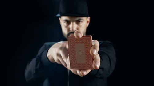 Professional Street Magician in a Cap Performs Sleight of Hand Card Tricks. Background is Black.