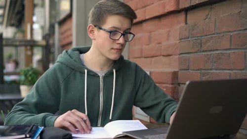 Young boy engaged in homework at cafe table with laptop and notepad in city