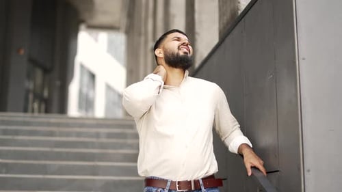 Tired adult handsome employee suffering from neck pain while standing on street near an office