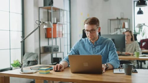 Handsome Young Man in Glasses and Shirt is Working on a Laptop in a Creative Business Agency. They