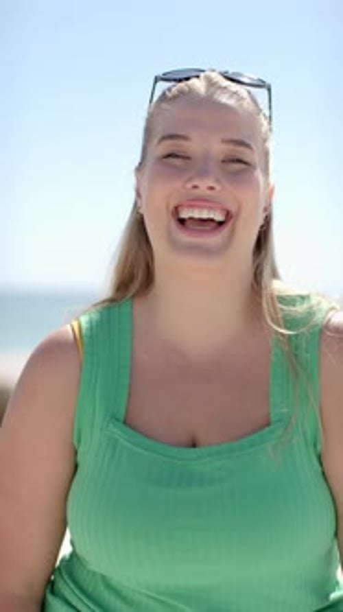 Vidéo verticale : Femme souriante profitant d'une journée ensoleillée à la plage, portant un haut vert et des lunettes de soleil