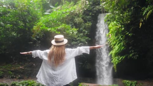 Carefree Blonde Woman in Straw Hat with Arms Spread Wide Enjoys Breathtaking Mountain Waterfall in