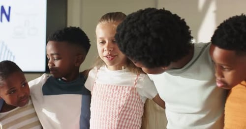 Diverse children linking arms, talking after display cue in class, girl in pink dress presenting