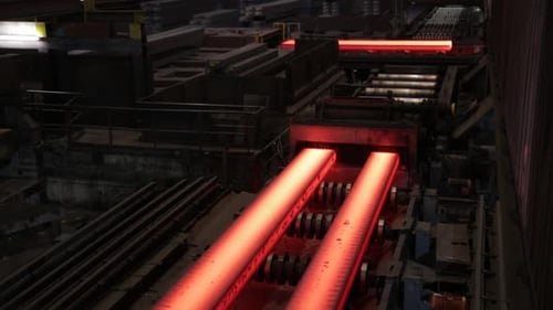 Glowing steel bars cooling on conveyor in a dark industrial setting, overhead view