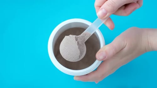 Chocolate protein powder in scoop and plastic jar on blue background, top view.