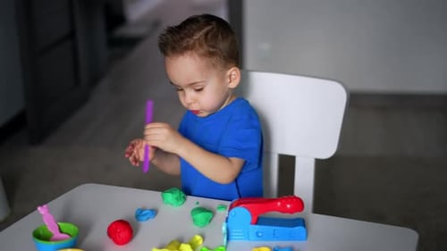 Small kid sits at desk busy with his hobby. Cute toddler plays with plasticine and stick. Top view.