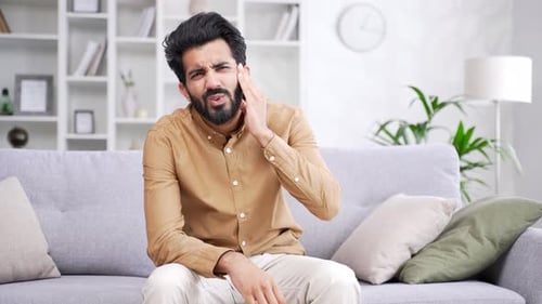 Earache. Sick young handsome man suffers from ear pain sitting on sofa at home. Upset male