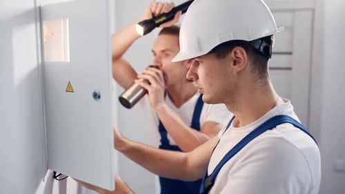Two young male electricians works indoors together. Using flashlight