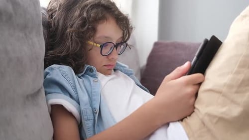 Little Cute Boy with Eyeglasses Sitting on Sofa and Playing Game on Digital Tablet