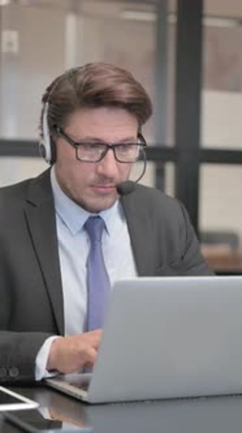 Call Center Man Looking at Camera while Working on Laptop with Headset, vertical