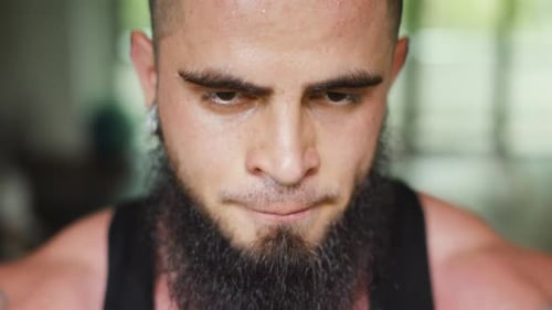Young man with beard looks into the camera, sweating and breathing strong after exercise in gym clos