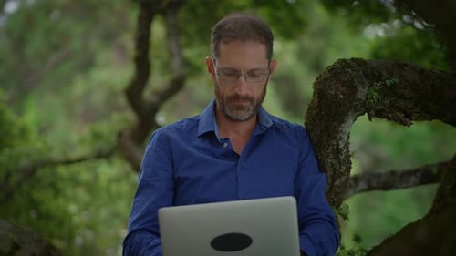 Man Works on Laptop Sitting on a Tree Branch
