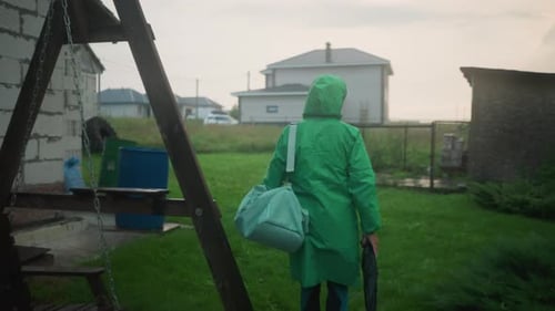 Individual in Green Raincoat Carrying Bag and Umbrella Walking Towards Grassy Area