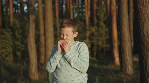 Caucasian Boy Bites Burger and Enjoys Taste in Park at Sunset