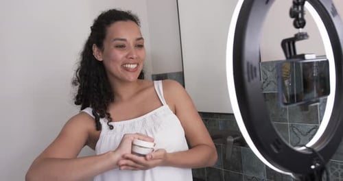 Mujer afroamericana filmando, probando la textura de un tarro, presentándolo a un teléfono inteligente en la luz del anillo del baño