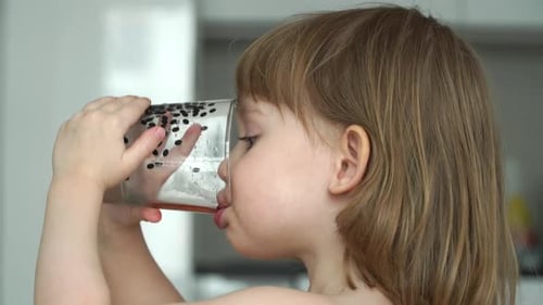 Une petite fille boit du jus de cerise dans un verre, regarde la caméra, rit et sourit