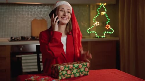 Young Woman in Santa Hat Talking on Phone While Wrapping Christmas Gifts at Home