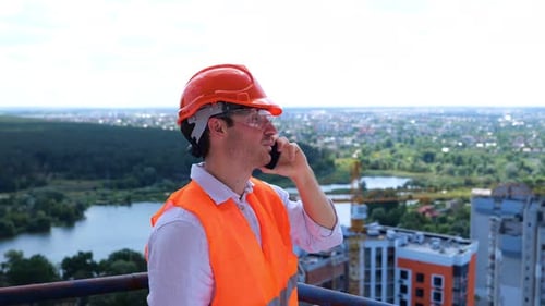 Smiling Young Builder Man in Helmet and Protective Glasses Talking on the Smartphone on the Roof
