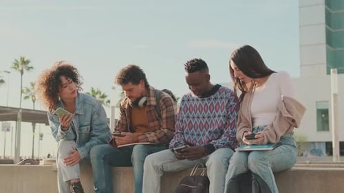 Students sit next to the university looking at their mobile phones and talking