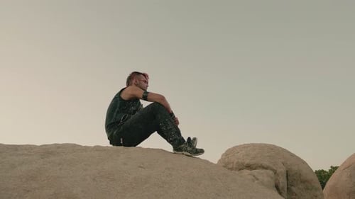 Punk style man sits atop a large rock against a pale sky.
