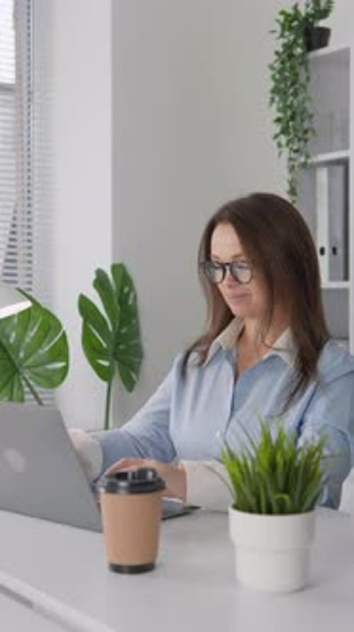 Une femme d'affaires est vue en train de travailler sur un ordinateur portable dans un bureau moderne entourée de café et de plantes