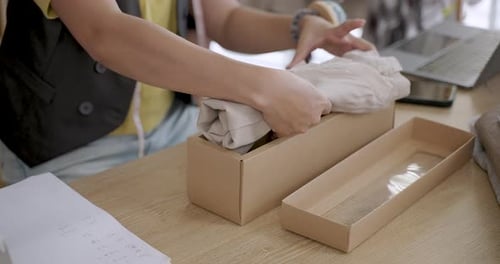 Asian female fashion business assistant placing clothing product inside cardboard box wrapping item