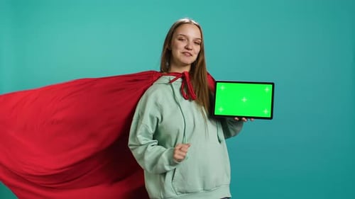 Joyeuse fille représentant un super-héros avec une cape rouge présentant une tablette à écran vert