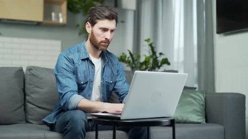 Serious handsome man works remotely at home with laptop while sitting on sofa. Confident male