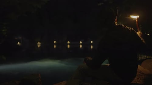 Man Sits with Torch at Night by Water