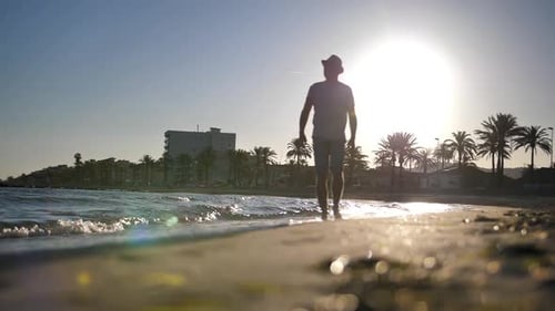 A Young Man in a Hat Walks Along the Beach During Sunset