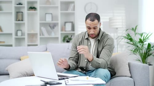 Müder junger Mann nimmt seine Brille ab und reibt sich die Augen, während er im Heimbüro am Laptop arbeitet