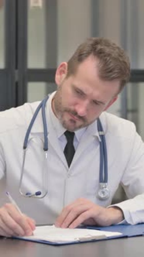 Male Doctor Writes on Clipboard at Desk Indoors